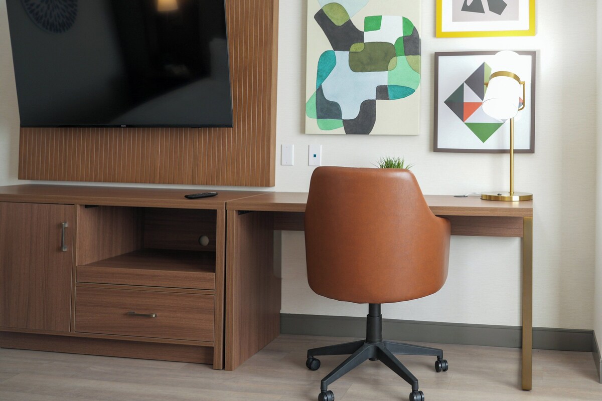 A modern office space with a wooden desk, a swivel chair, a flat-screen TV mounted on the wall, and abstract artwork on the wall.