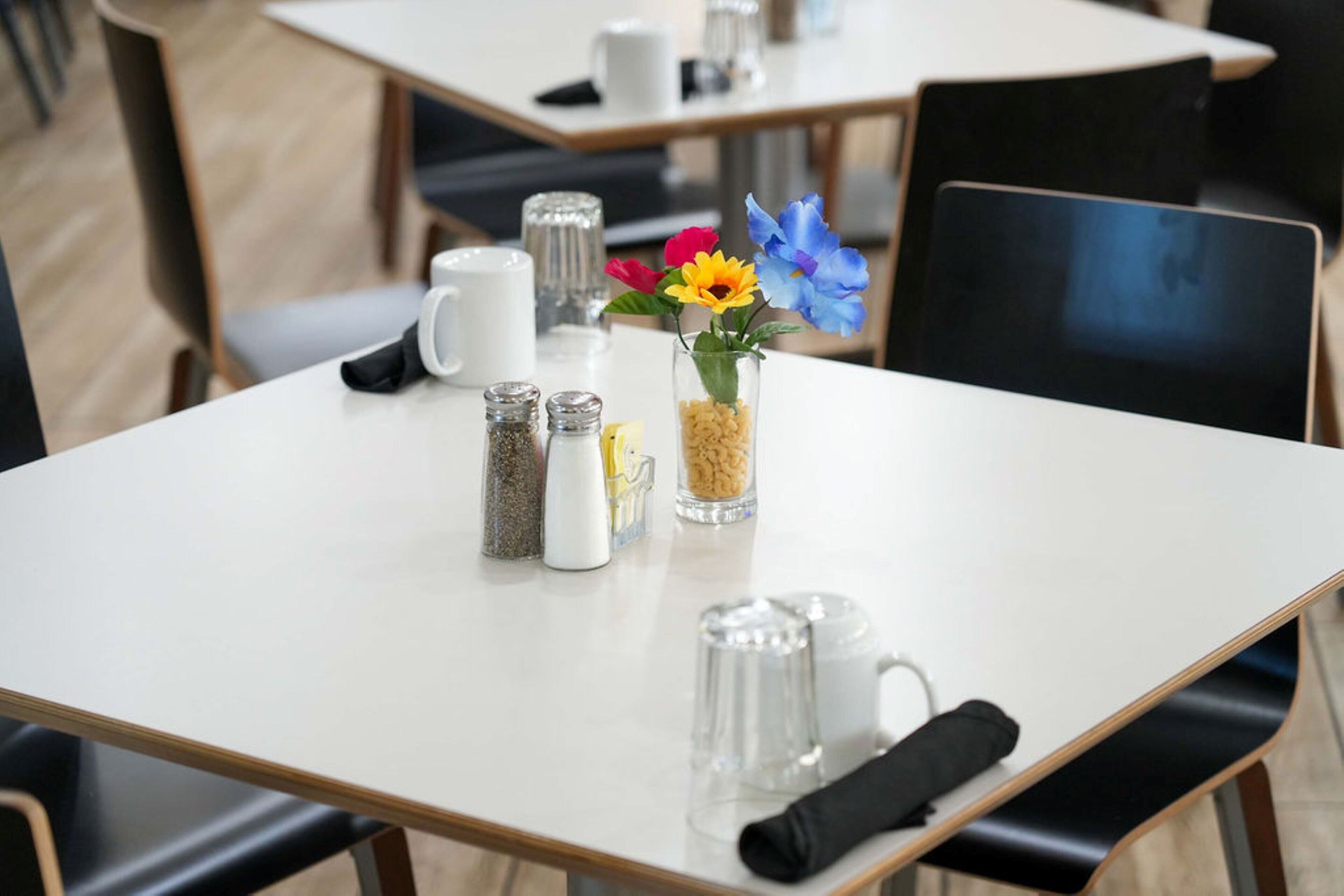 Un comedor limpio y moderno con una mesa blanca dispuesta para dos personas, que incluye saleros y pimenteros, un pequeño jarrón con flores y una jarra de vidrio transparente.