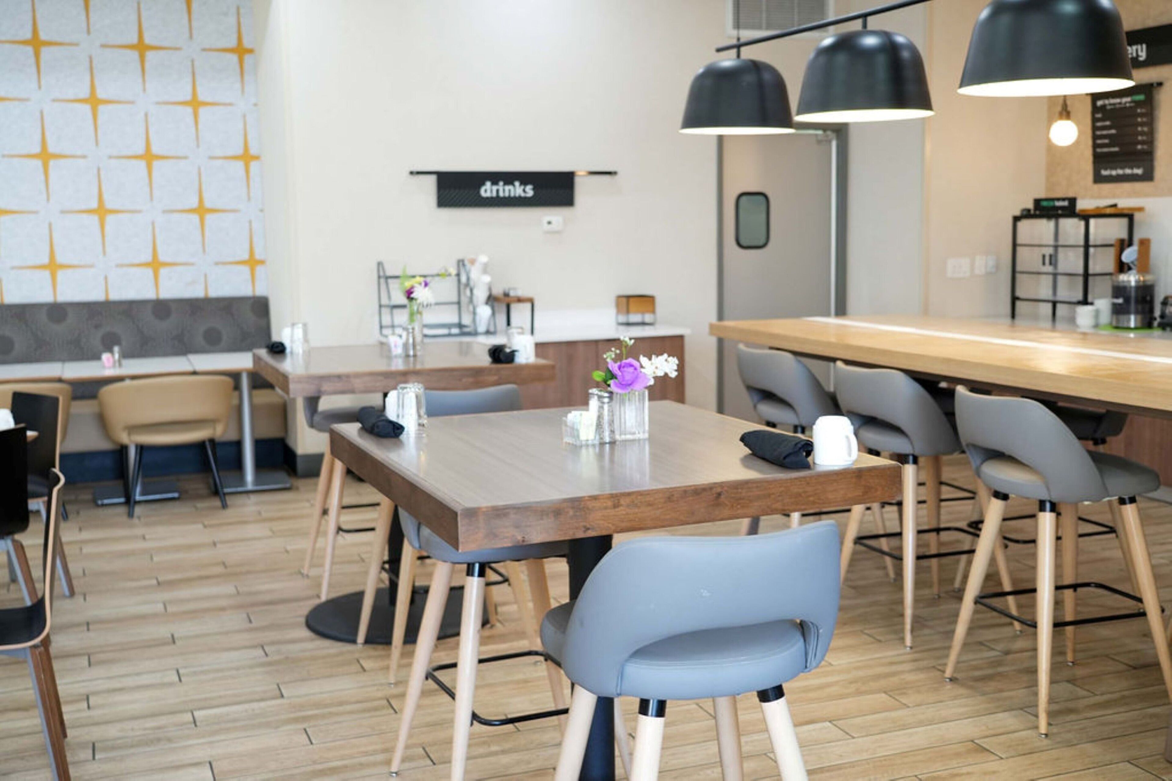Un café moderno con mesas y sillas de madera, una zona de bar y un letrero que dice 'drinks' en la pared.