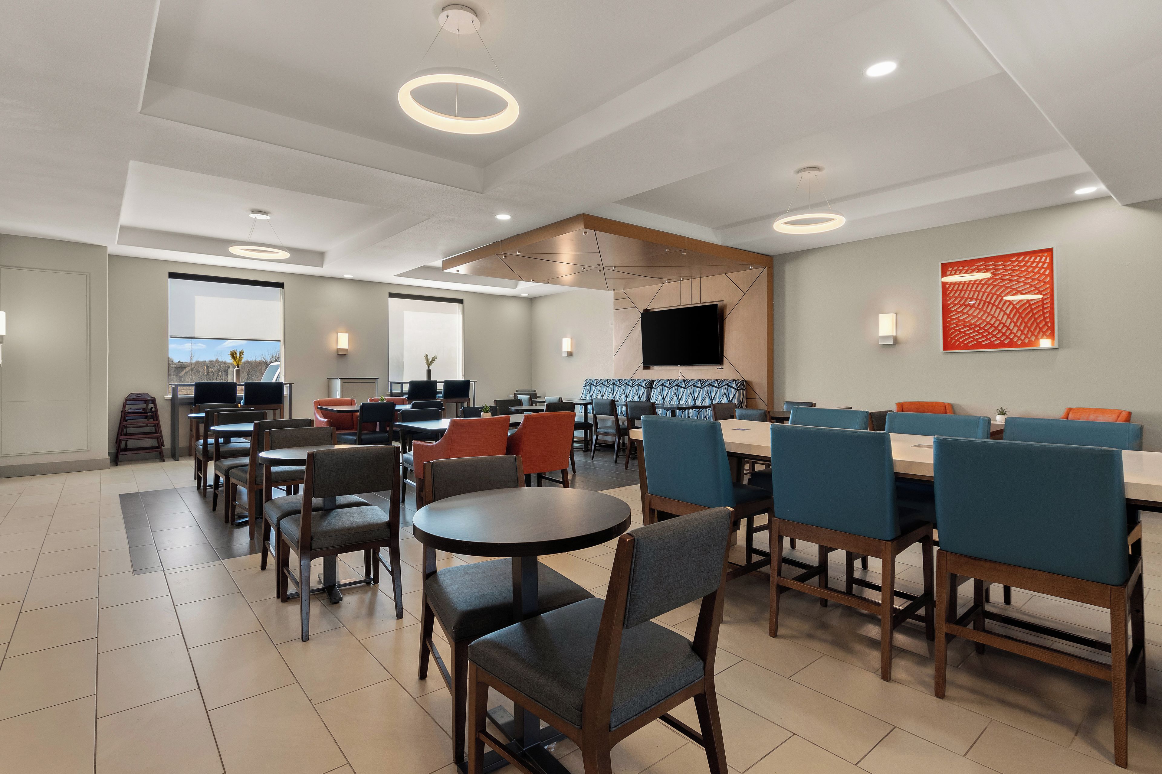 A modern dining area with round tables and chairs, recessed ceiling lighting, and a flat-screen TV mounted on the wall.