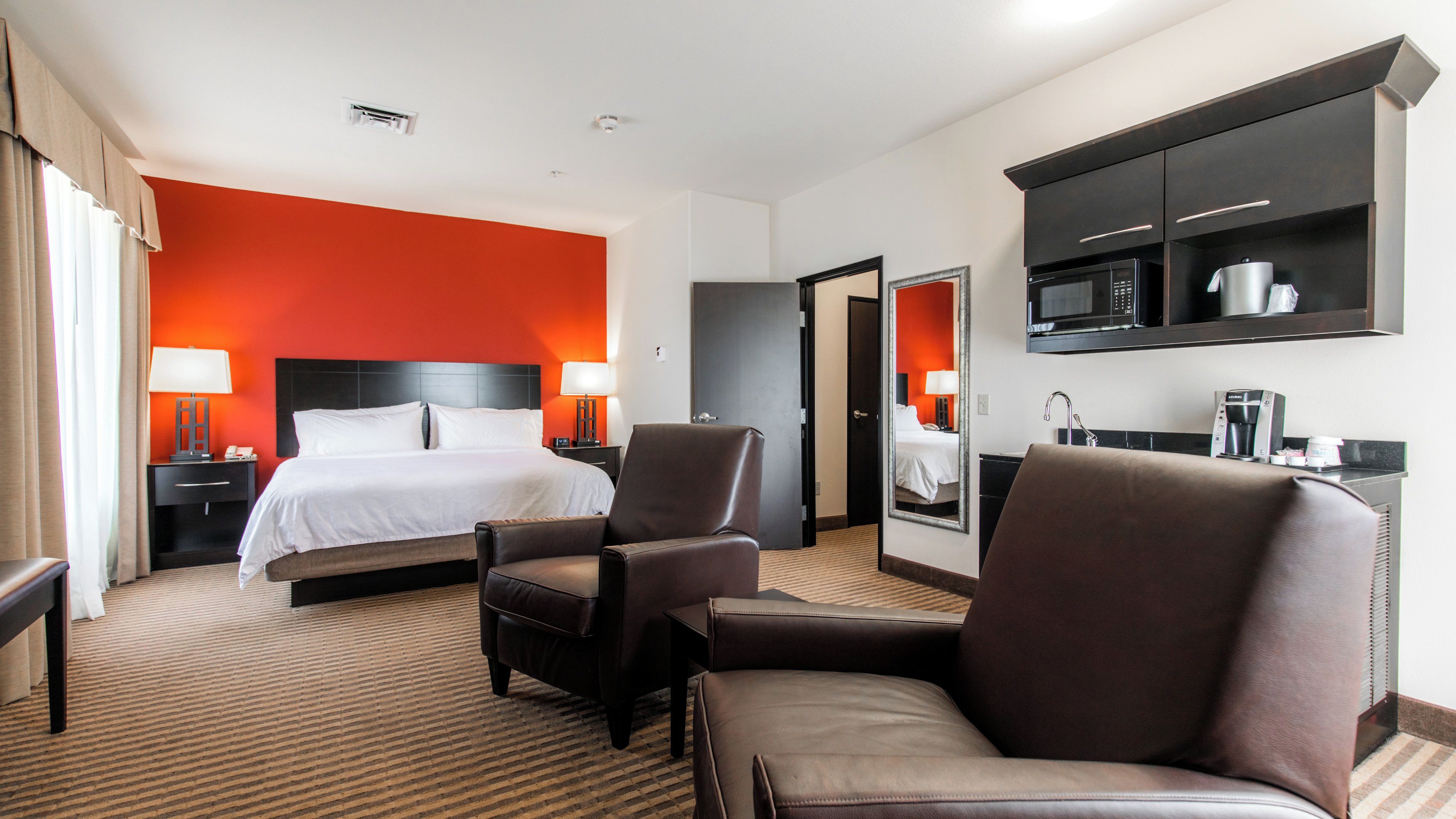 Habitación de hotel con cama, dos sillones, un escritorio con lámpara, minibar y espejo.