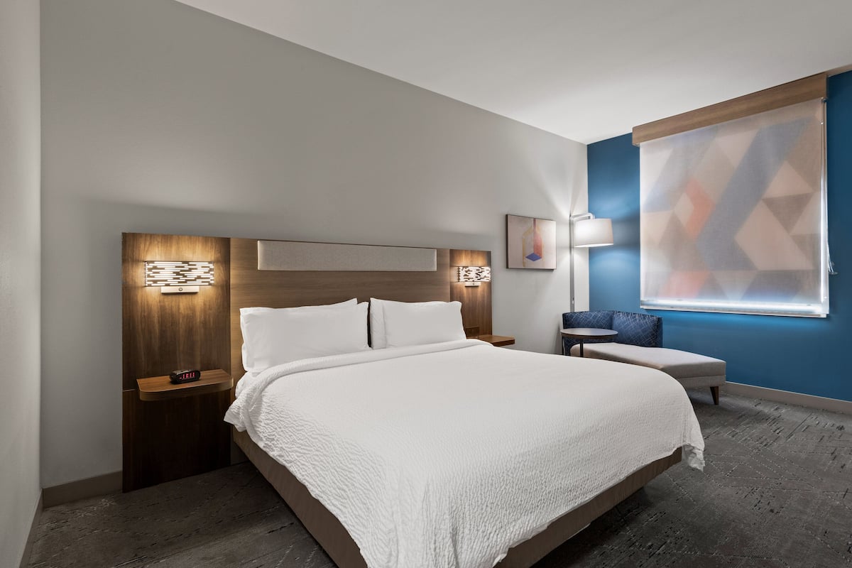 Moderni hotellihuone, jossa on suuri sänky, yöpöydät, yövalaisin, pieni pöytä, tuoli ja suuri ikkuna, jossa on geometriset sälekaihtimet.