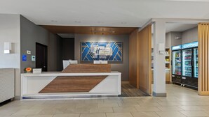A hotel reception area with a modern design, including a reception desk, a wall-mounted sign, and a refrigerator stocked with beverages. - Holiday Inn Express & Suites Oklahoma City NW-Quail Springs by IHG (Oklahoma City)