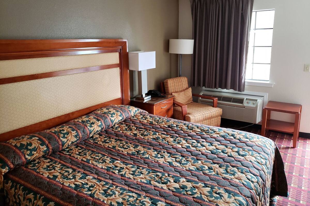 A hotel room with a large bed, a chair, a lamp, an air conditioner, and a window with curtains.