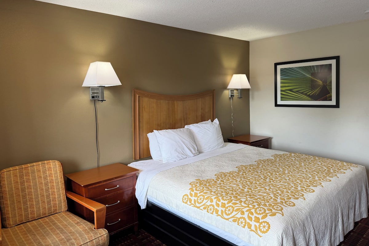 A hotel room with a bed, two bedside lamps, a chair, a nightstand, and a framed picture on the wall.