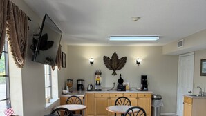 Un comedor limpio y bien iluminado con mesas y sillas redondas, una cocina al fondo y una hoja decorativa en la pared.
