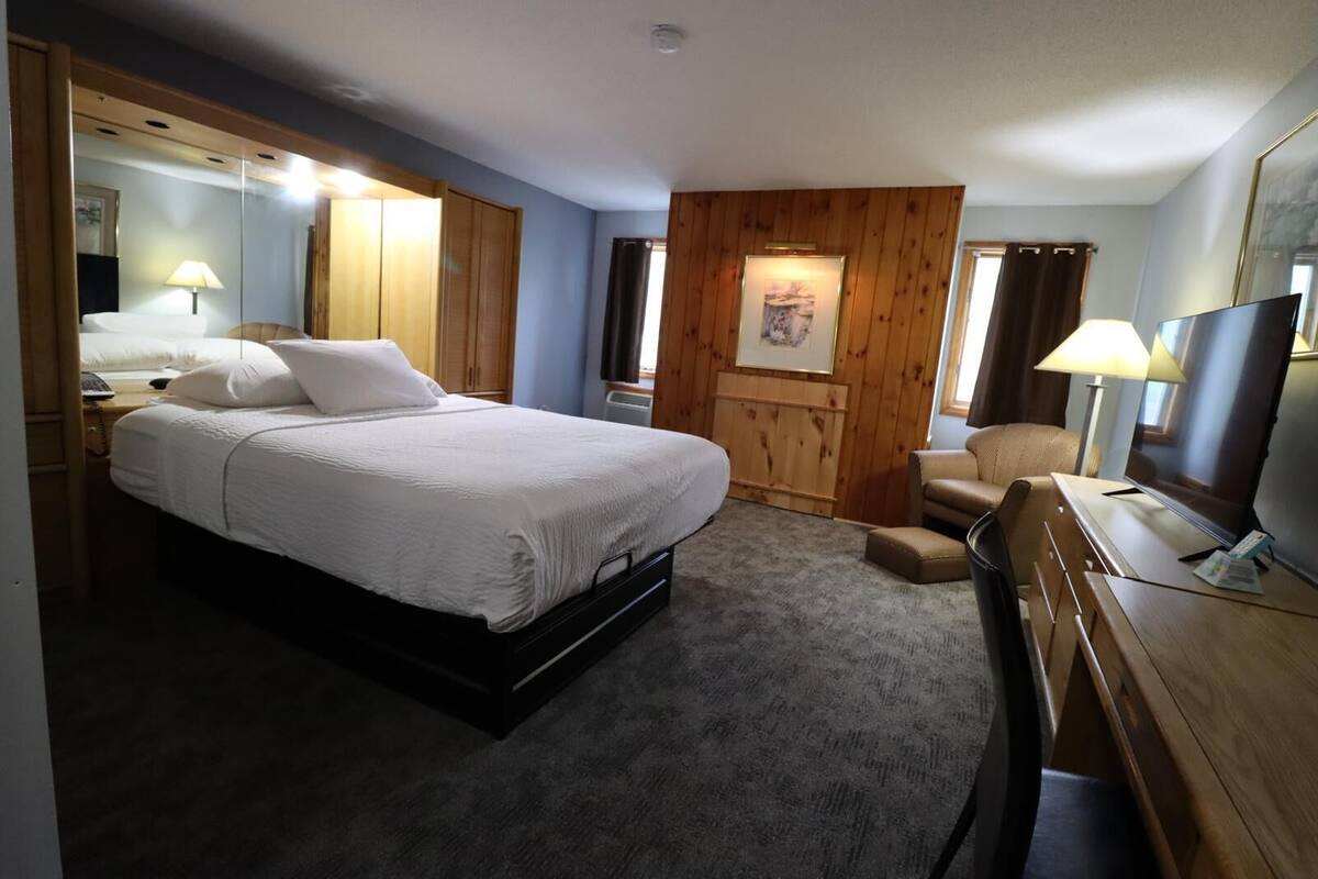 Ein Hotelzimmer mit einem großen Bett, einem Schreibtisch mit Fernseher, einem Sessel und einer Wand mit Holzvertäfelung.
