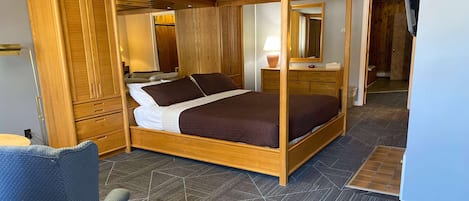 A bedroom with a four-poster bed, wooden wardrobe, bedside table, lamp, and a mirror.