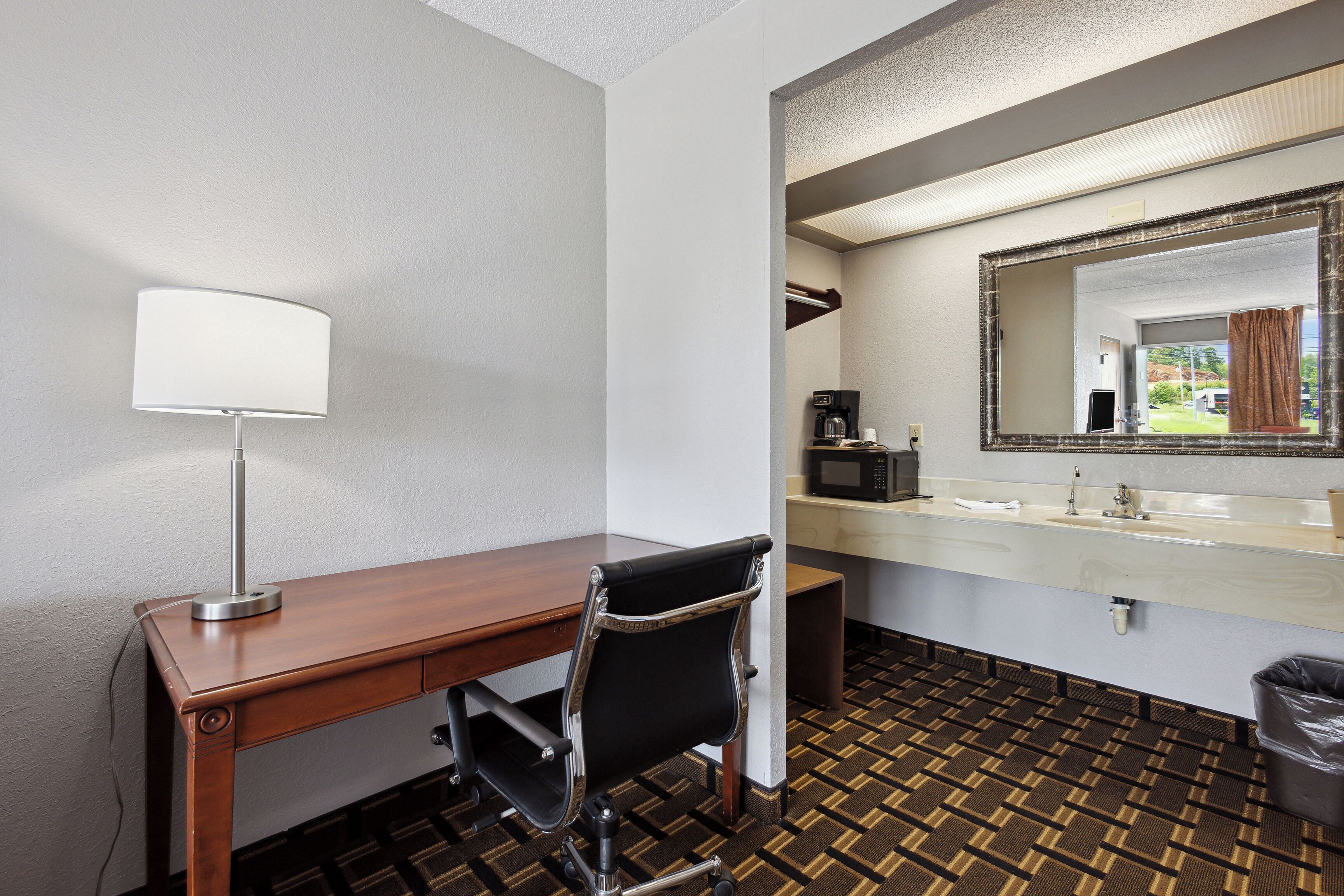 Habitación de hotel con un escritorio de madera, una silla de oficina negra, una lámpara blanca, un microondas, un lavabo y un suelo estampado.