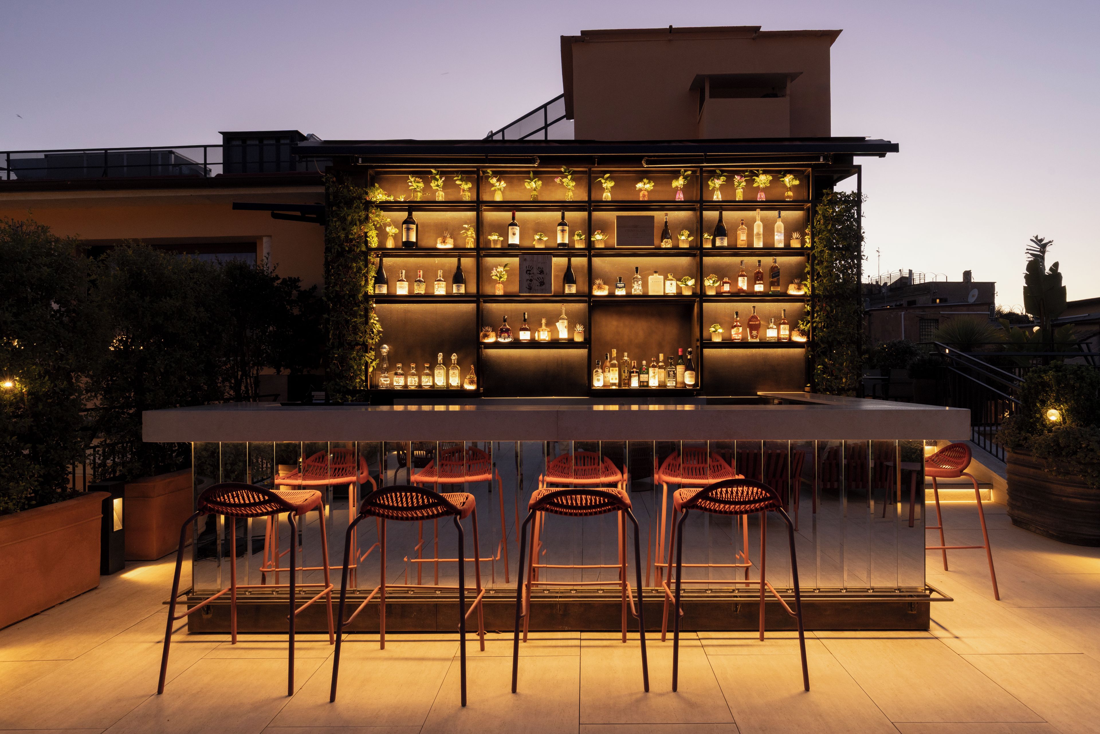 An outdoor bar area with multiple shelves of bottles, a seating arrangement with chairs, and a backdrop of buildings.
