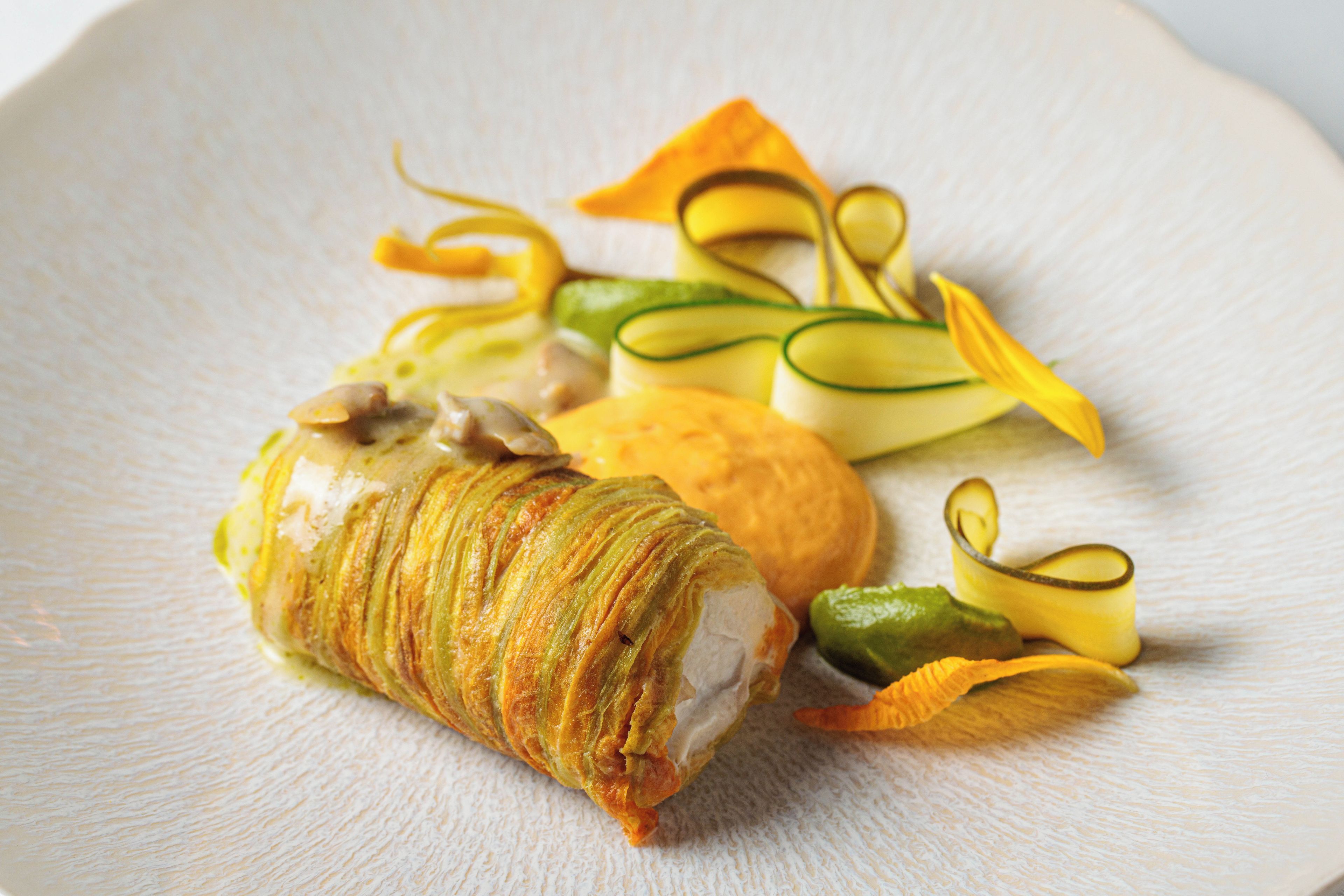 Une assiette contenant un amuse-gueule enroulé, un petit pain rond et quelques légumes en guise de garniture.