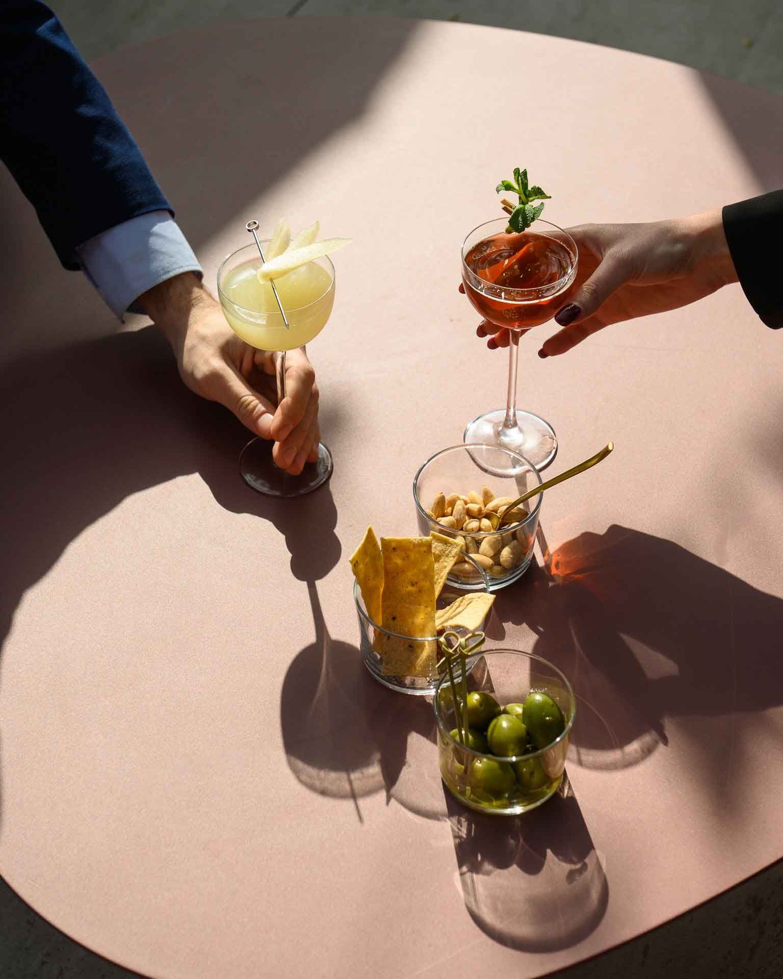 A table with two drinks, a bowl of peanuts, and a bowl of olives.