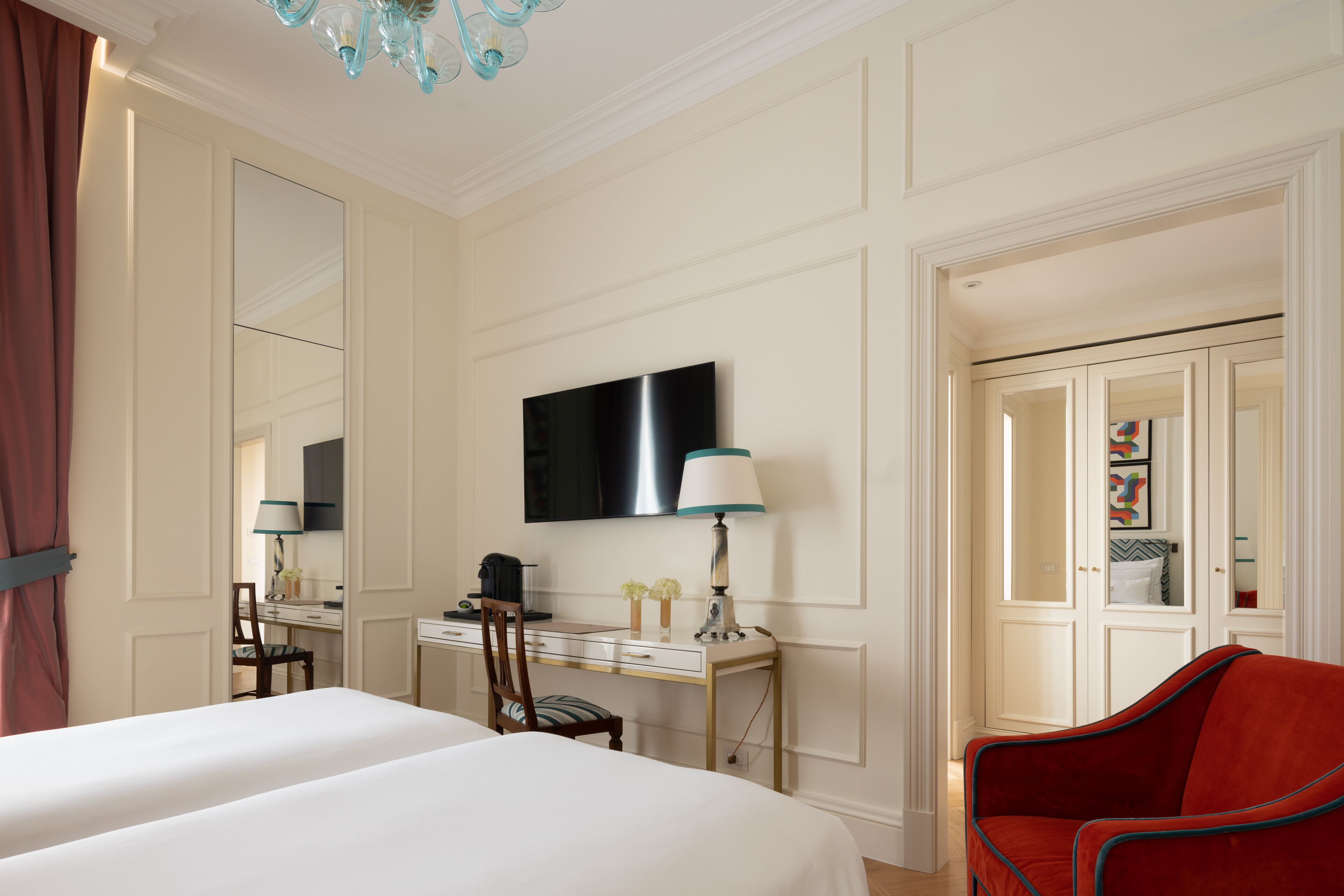 A hotel room with a bed, desk, chair, TV, and a red armchair.