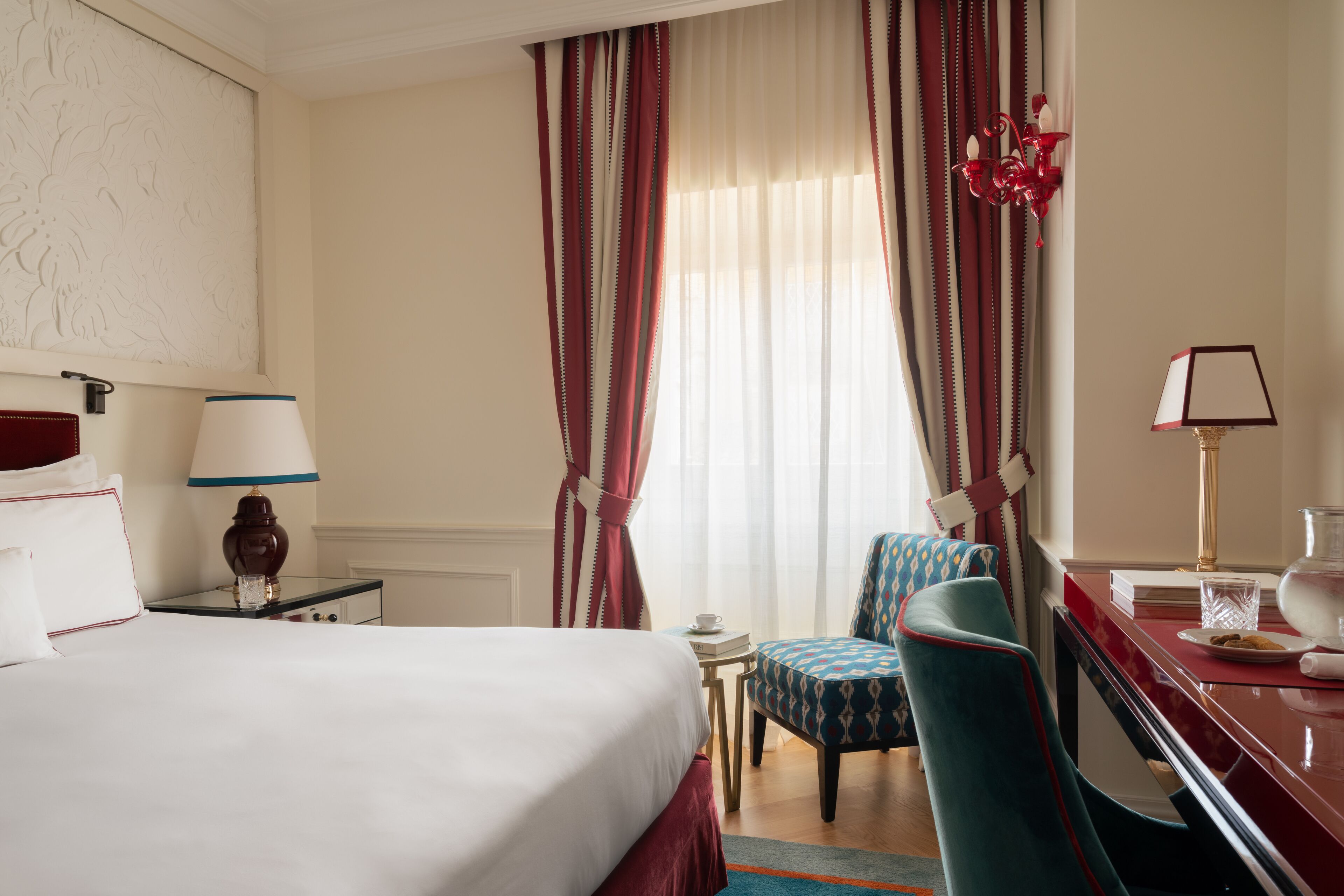 Une chambre d’hôtel avec un lit, un bureau avec une lampe, une chaise, une petite table avec une tasse et des rideaux.