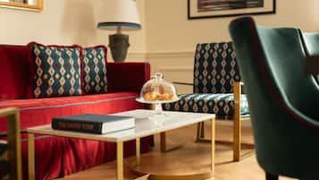 Un salon avec un canapé rouge, des coussins à motifs, un fauteuil turquoise, une table basse sur laquelle repose un livre intitulé « Le Grand Tour », et une lampe.