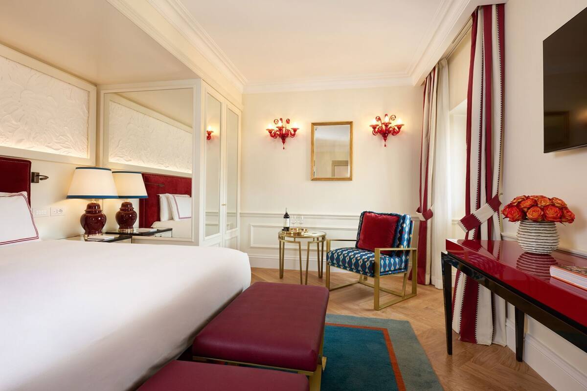 A hotel room with a bed, a chair, a small table with a bottle and glasses, a mirror, and a vase with flowers.
