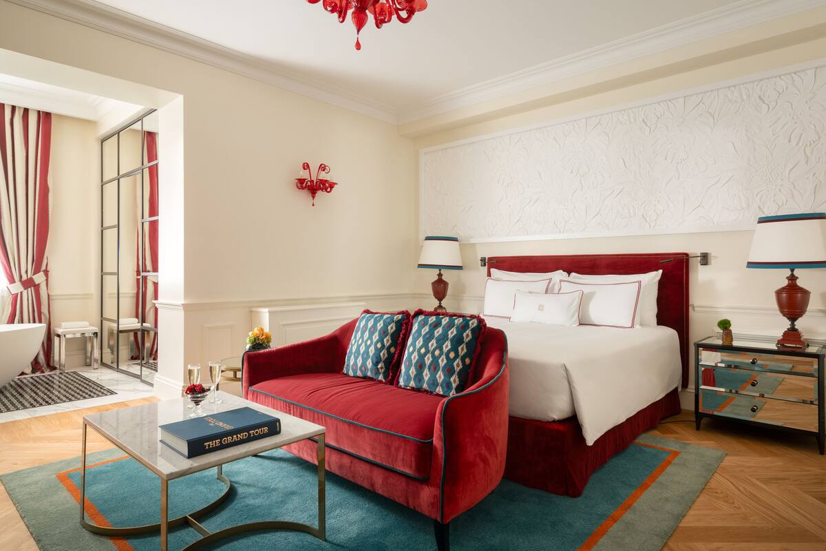 A hotel room with a red sofa, a bed with white bedding, a small table with a book, and a nightstand with a lamp.