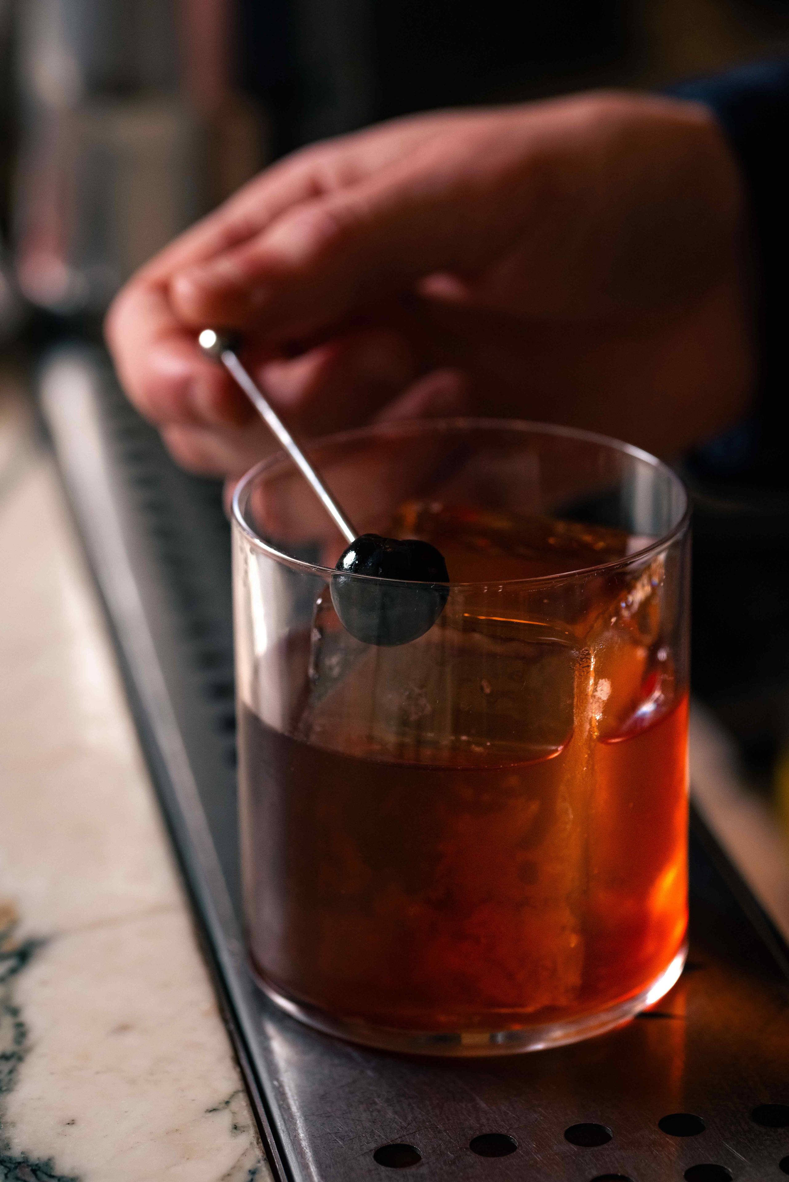 Um copo de vidro transparente com um líquido escuro, possivelmente um drink, e uma colher apoiada na borda.