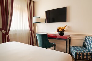 Camera d'albergo con un letto, una scrivania con un vaso di fiori, una sedia, un televisore a parete e tende.