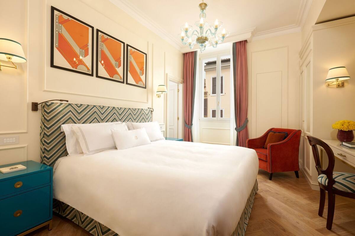 A hotel room with a bed, a nightstand, a chair, a desk, and a chandelier.