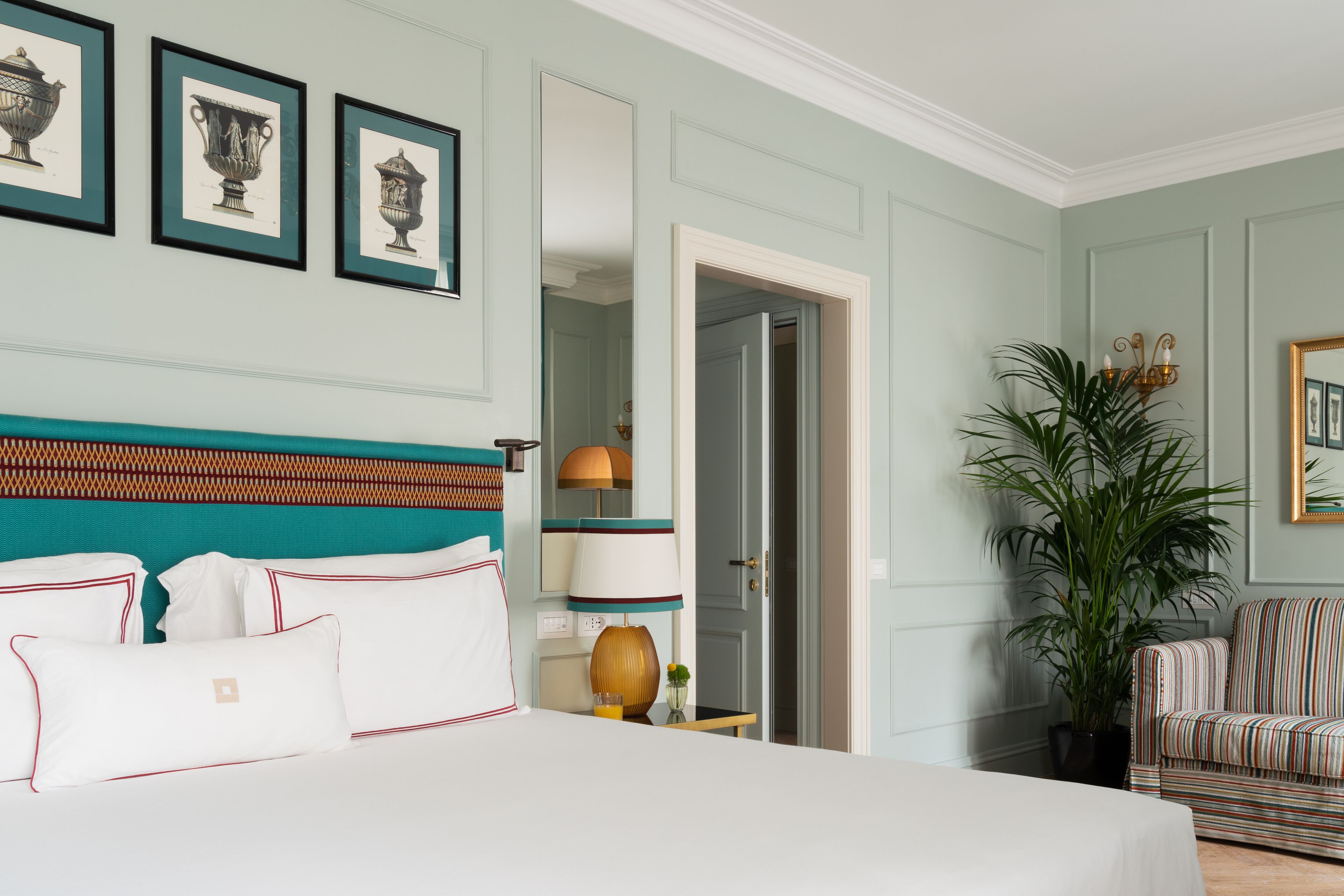 Una habitación de hotel con una cama grande, una mesita de noche con lámpara, una planta en maceta y un sillón a rayas.