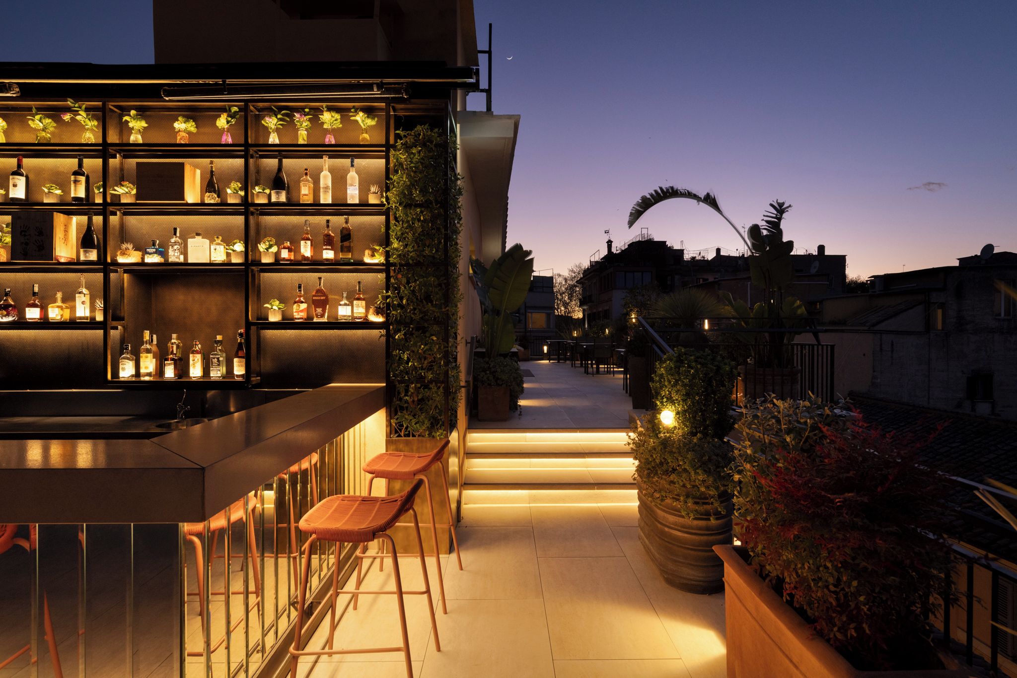 An outdoor bar area with shelves of bottles, a seating area with stools, and a view of the cityscape at dusk.