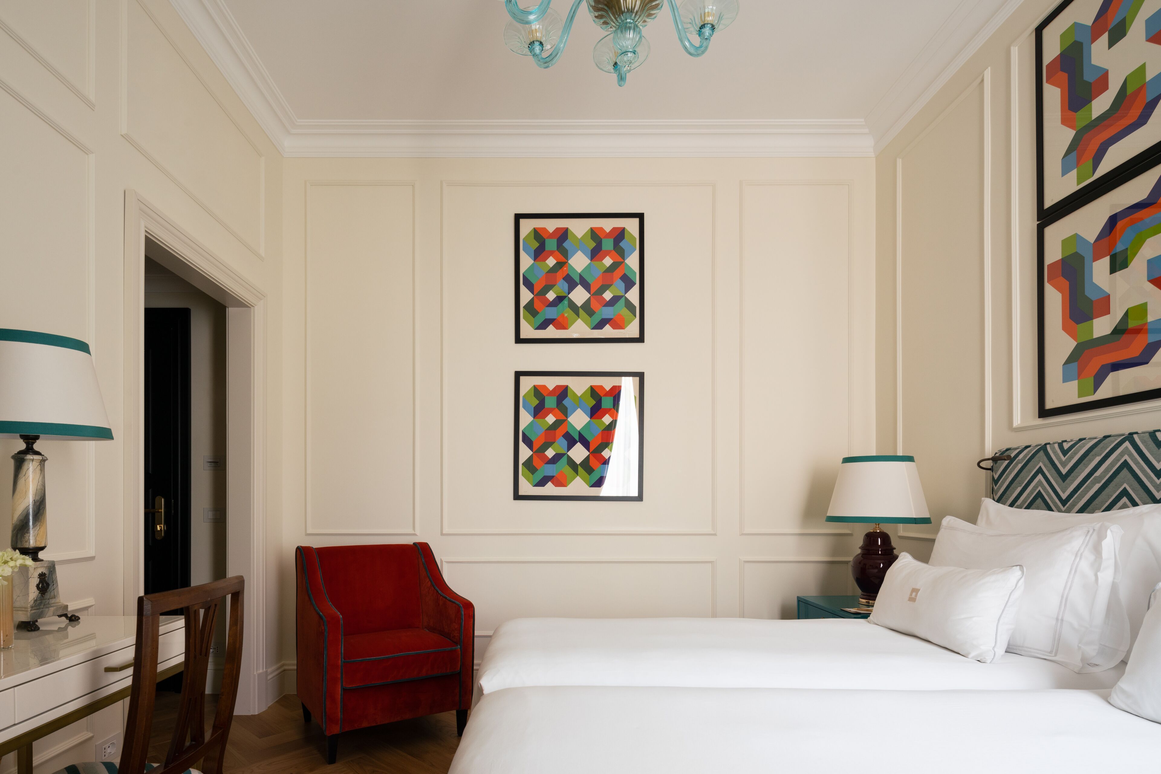 Een hotelkamer met een bed, een rode stoel, een bureau met een lamp en twee kunstwerken in lijsten aan de muur.