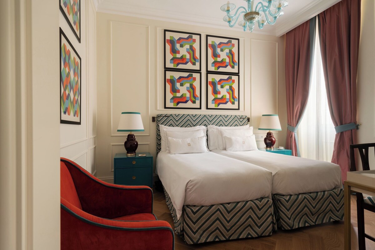 Quarto de hotel com duas camas, uma poltrona vermelha e obras de arte nas paredes.