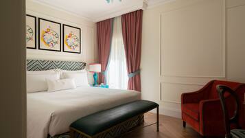 A hotel room with a bed, a bench, a red armchair, a nightstand with a lamp, and a window with curtains.