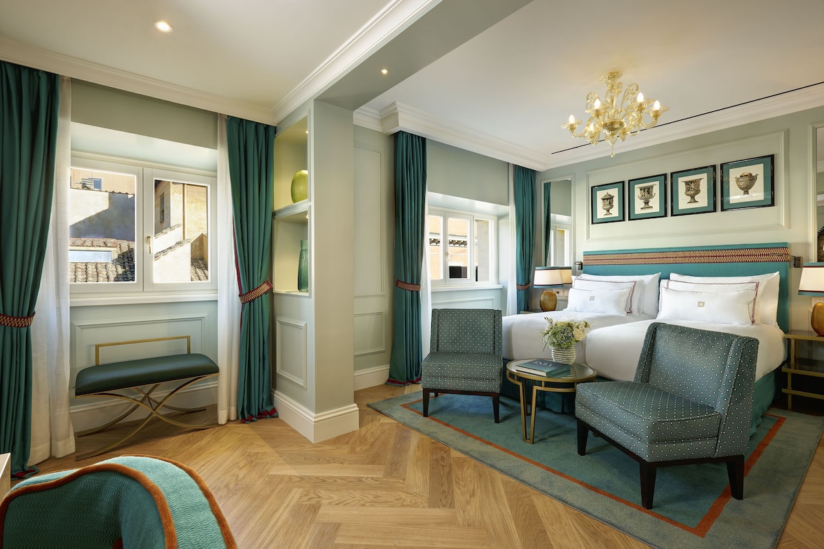 A hotel room with a bed, two armchairs, a small table, a chandelier, and a view of the outside.