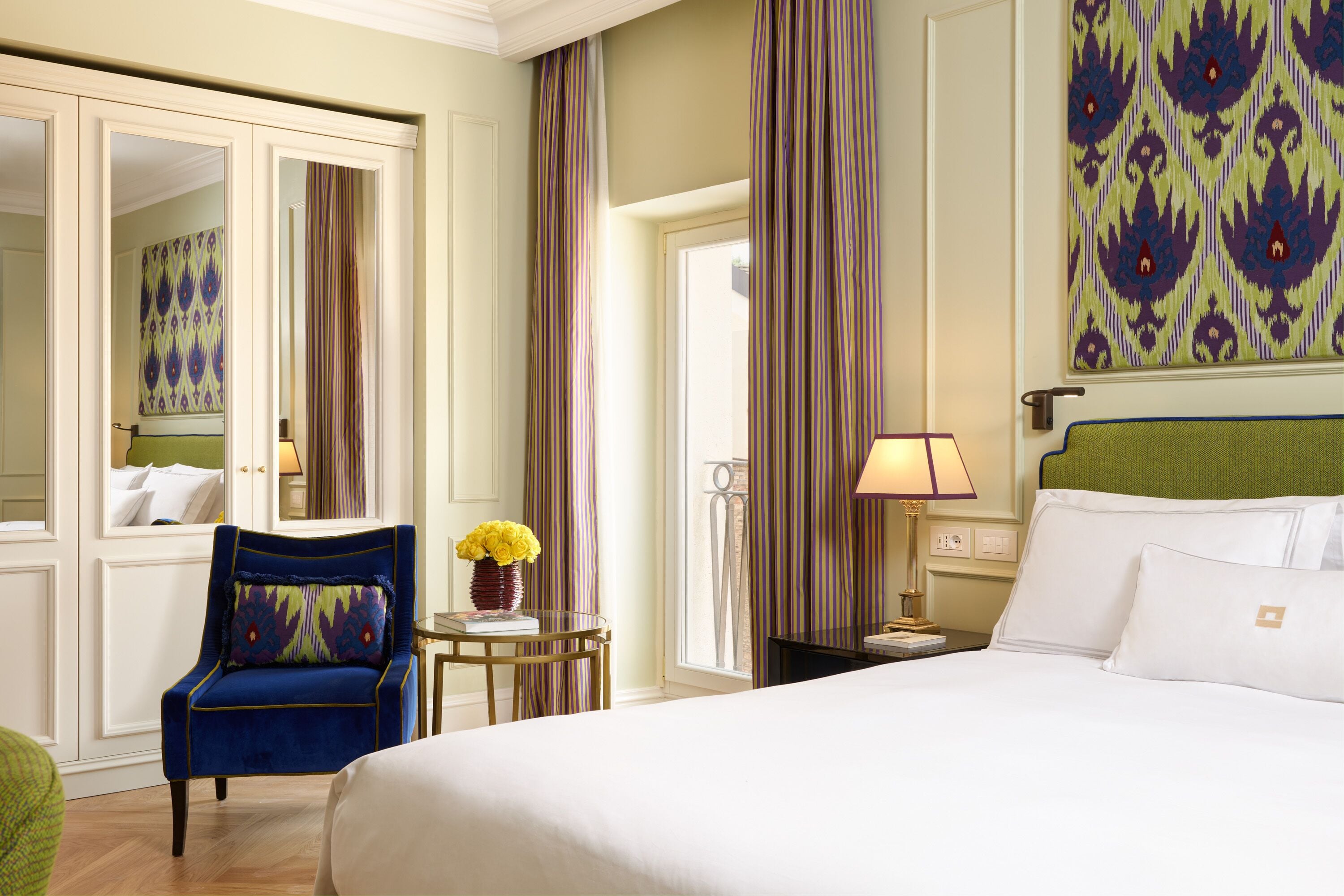 A hotel room with a large bed, a blue armchair, a small table with a vase of flowers, and a view of the outdoors through a window.