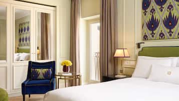 A hotel room with a large bed, a blue armchair, a small table with a vase of flowers, and a view of the outdoors through a window.