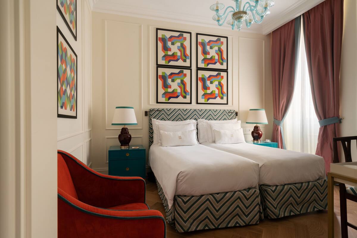 A hotel room with two beds, a red armchair, a nightstand with a lamp, and a wall with four framed artworks.