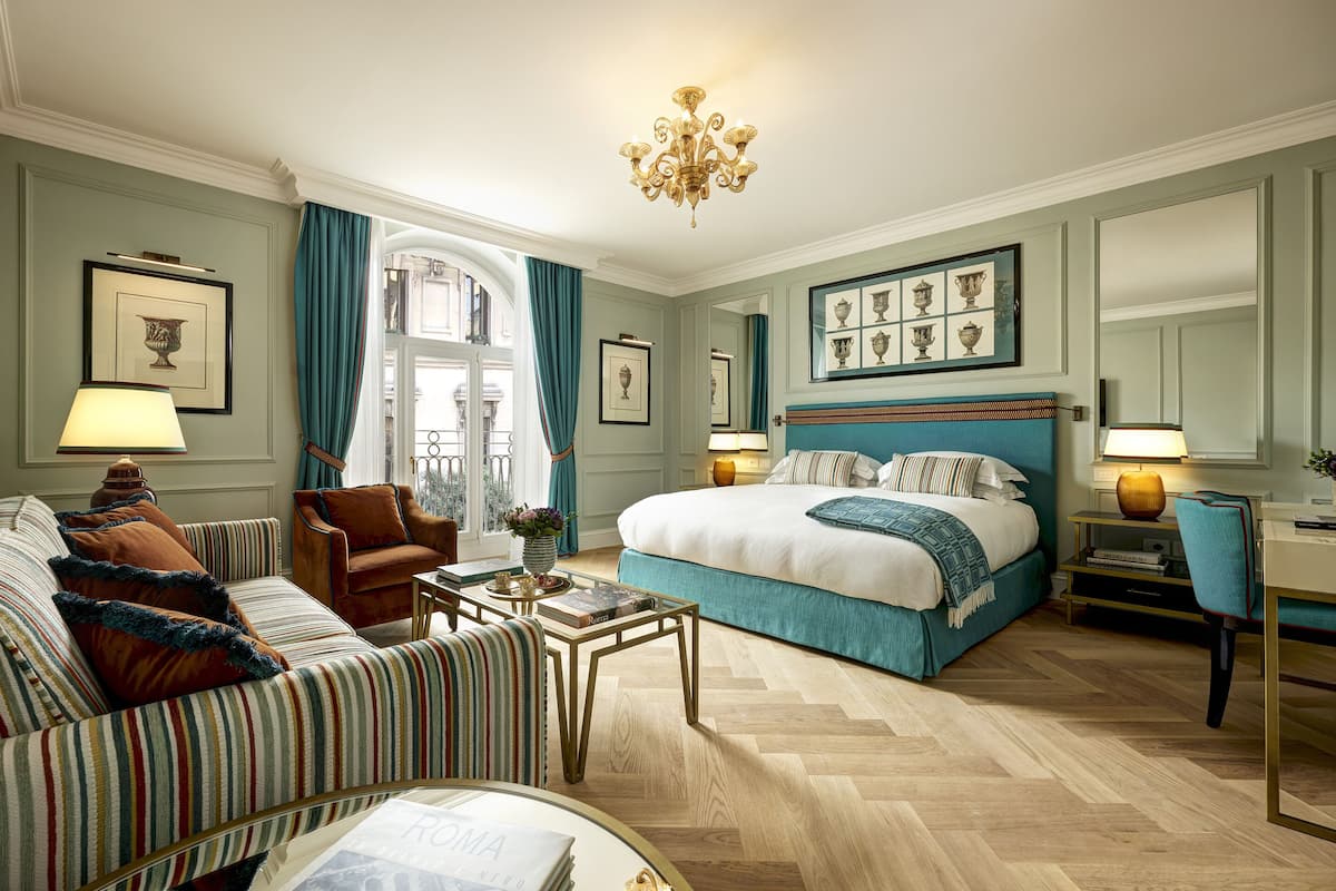 A well-appointed hotel room with a large bed, a sitting area with a sofa and armchair, a desk with a chair, and a chandelier.