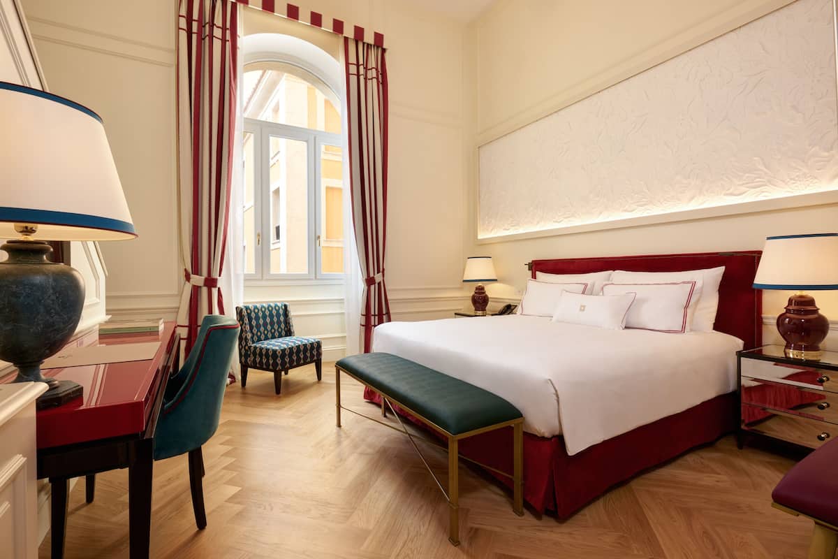 A hotel room with a large bed, a desk with a lamp, a chair, a bench, and a window with curtains.