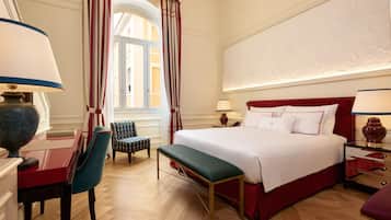 A hotel room with a large bed, a desk with a lamp, a chair, a bench, and a window with curtains.