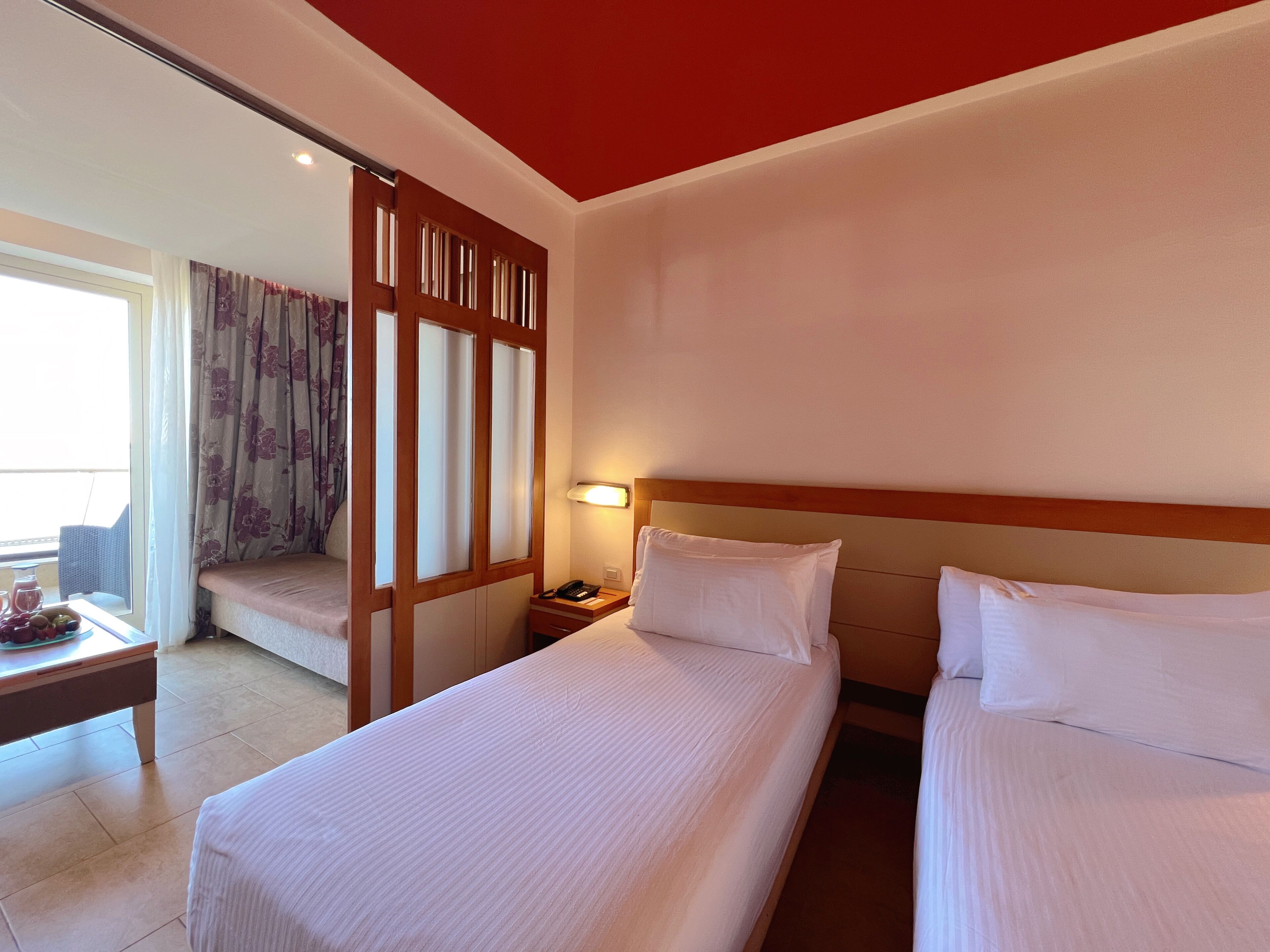 A hotel room with two beds, a wooden partition, a bedside table with a lamp, and a view of the balcony.