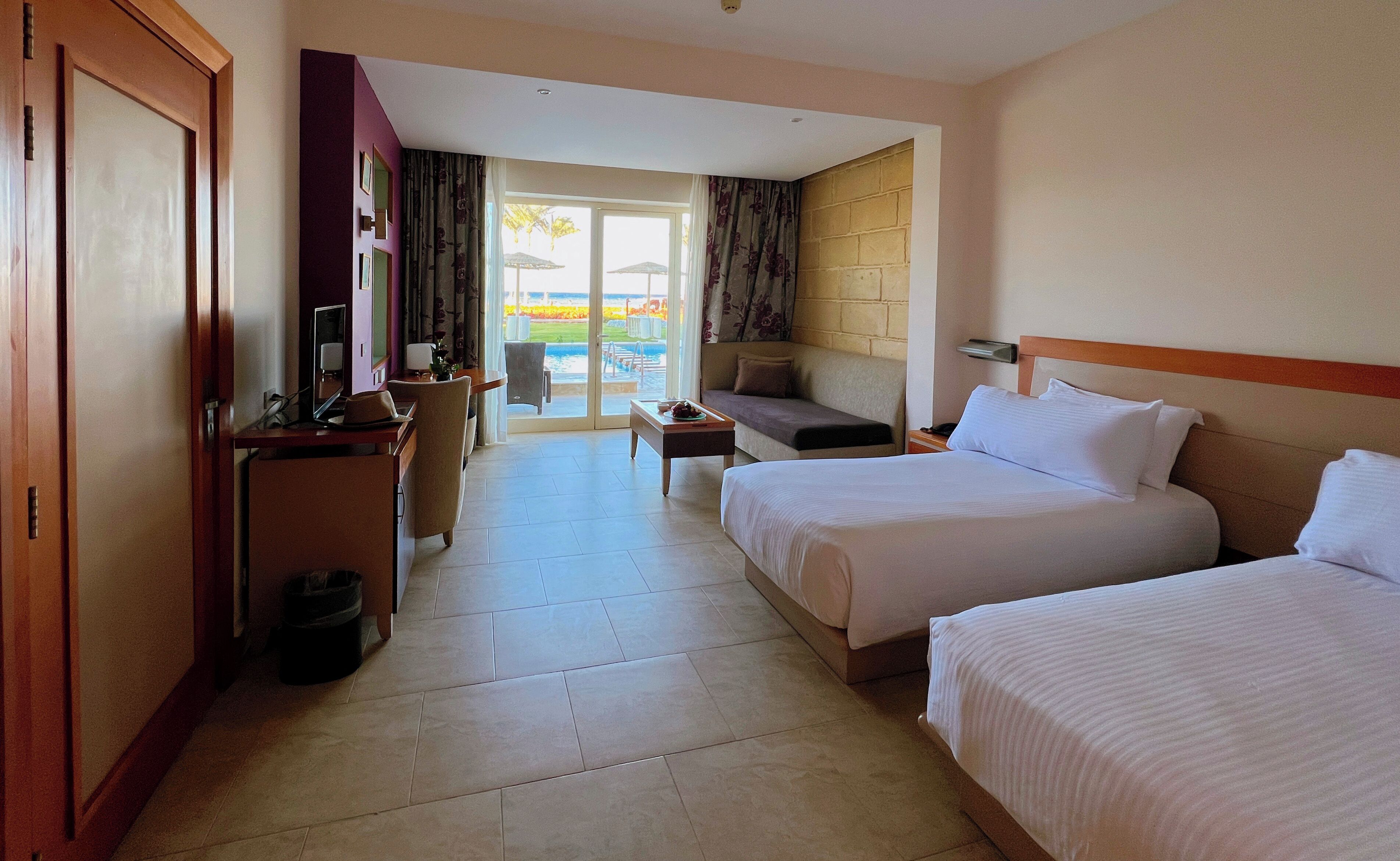 Habitación de hotel con dos camas, un escritorio con computadora, una silla y un sofá. Se observa una vista de la piscina y palmeras a través de la puerta de cristal.