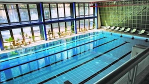 An indoor swimming pool with clear lanes, a sunlit area with chairs and plants, and a view through large windows. - Hotel Samokov (Samokov)