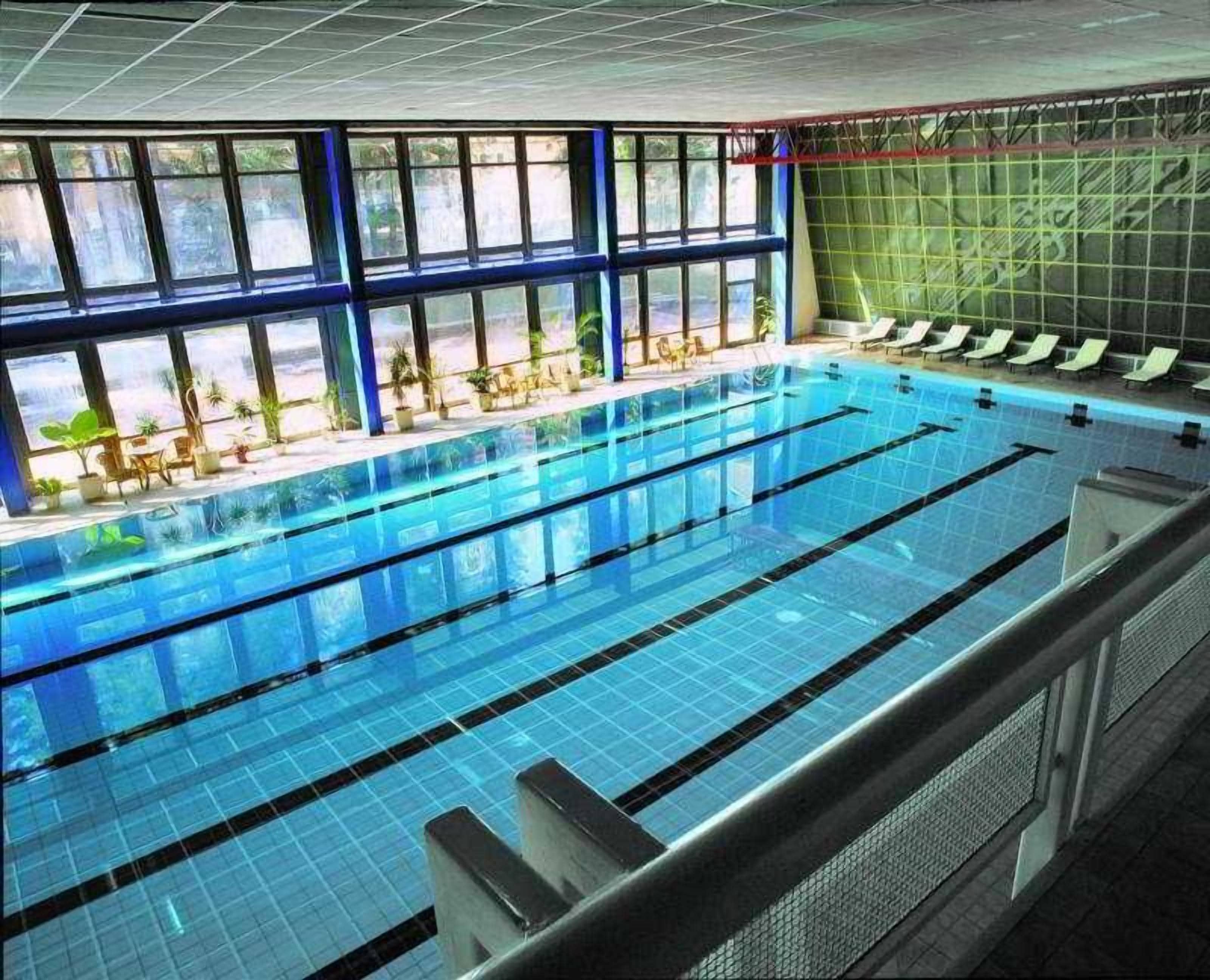 An indoor swimming pool with multiple lanes, a sunlit area with chairs, and a view of the outdoors through large windows.