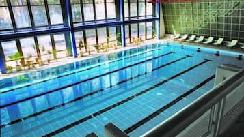 An indoor swimming pool with multiple lanes, a sunlit area with chairs, and a view of the outdoors through large windows.