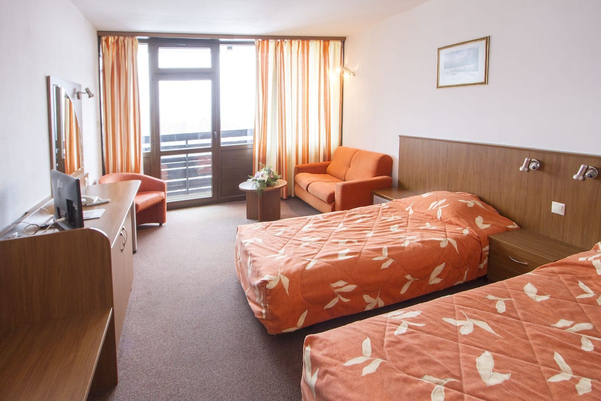 A hotel room with two beds, a desk, a chair, a sofa, and a balcony with curtains.