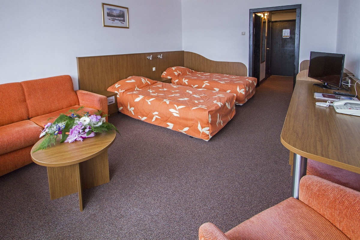 A hotel room with a wooden bed, a wooden desk, a wooden chair, a sofa, and a small table with flowers.