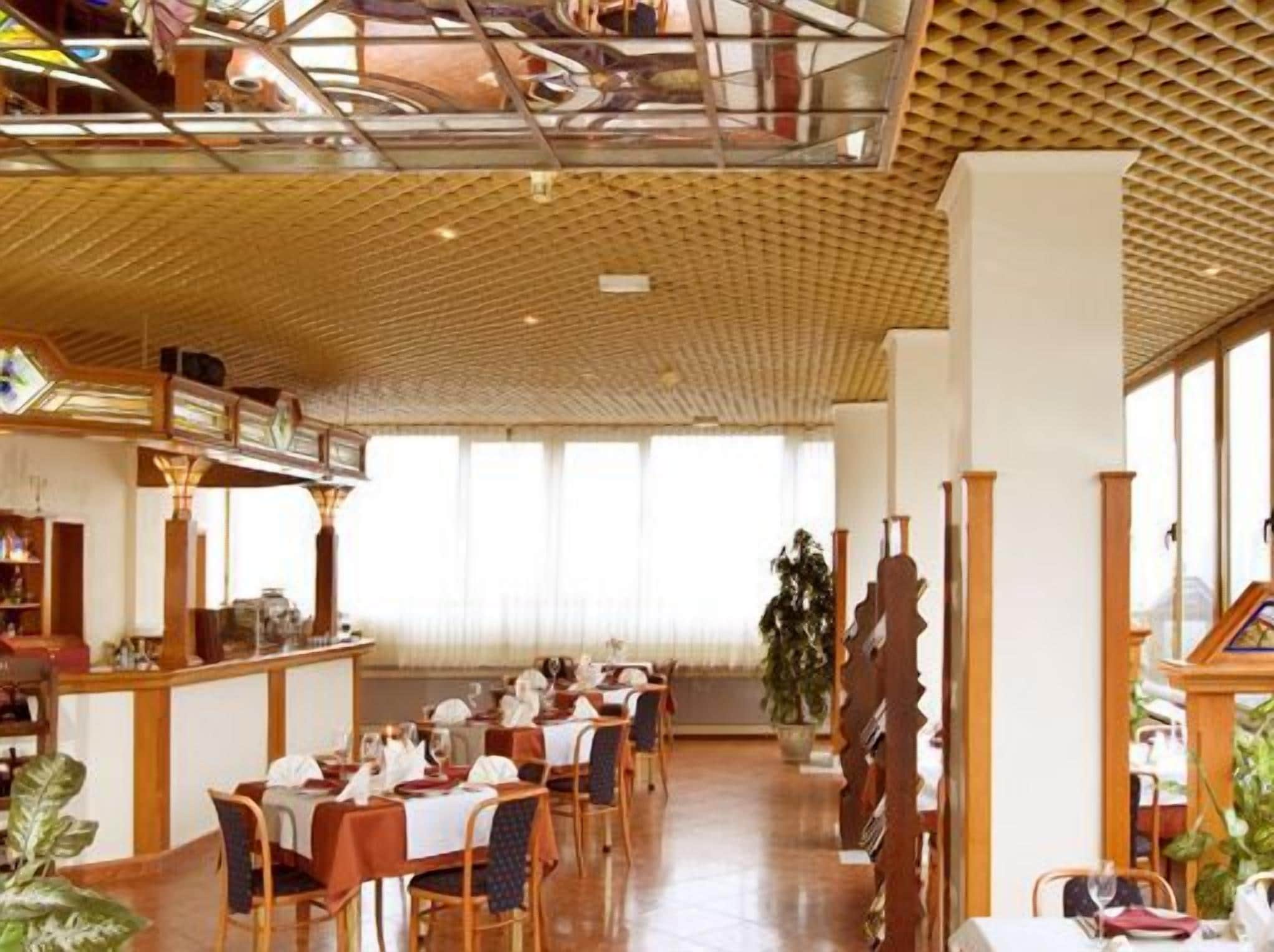 A restaurant with a high ceiling, wooden beams, and large windows allowing natural light. Tables are set with white tablecloths and chairs.