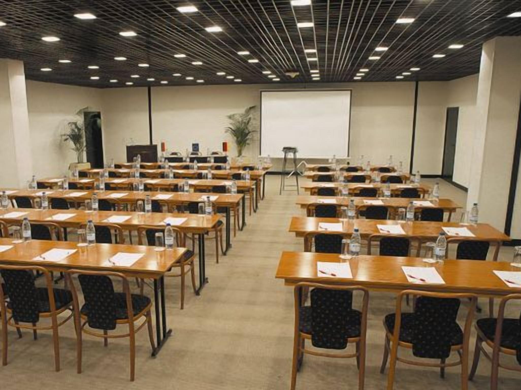 A conference room with rows of tables and chairs, equipped with water bottles and papers, and a projector screen at the front.