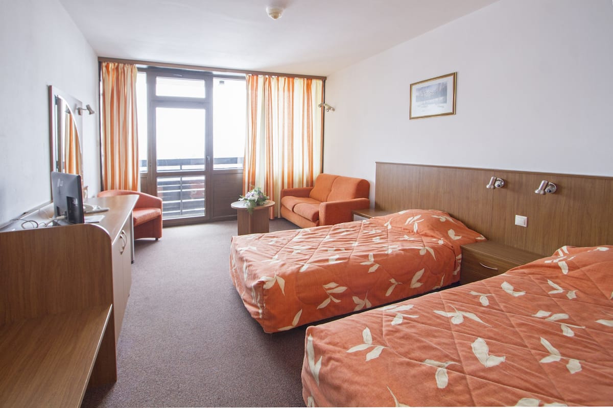 A hotel room with two beds, a desk, a chair, a small table with a plant, and a window with curtains.