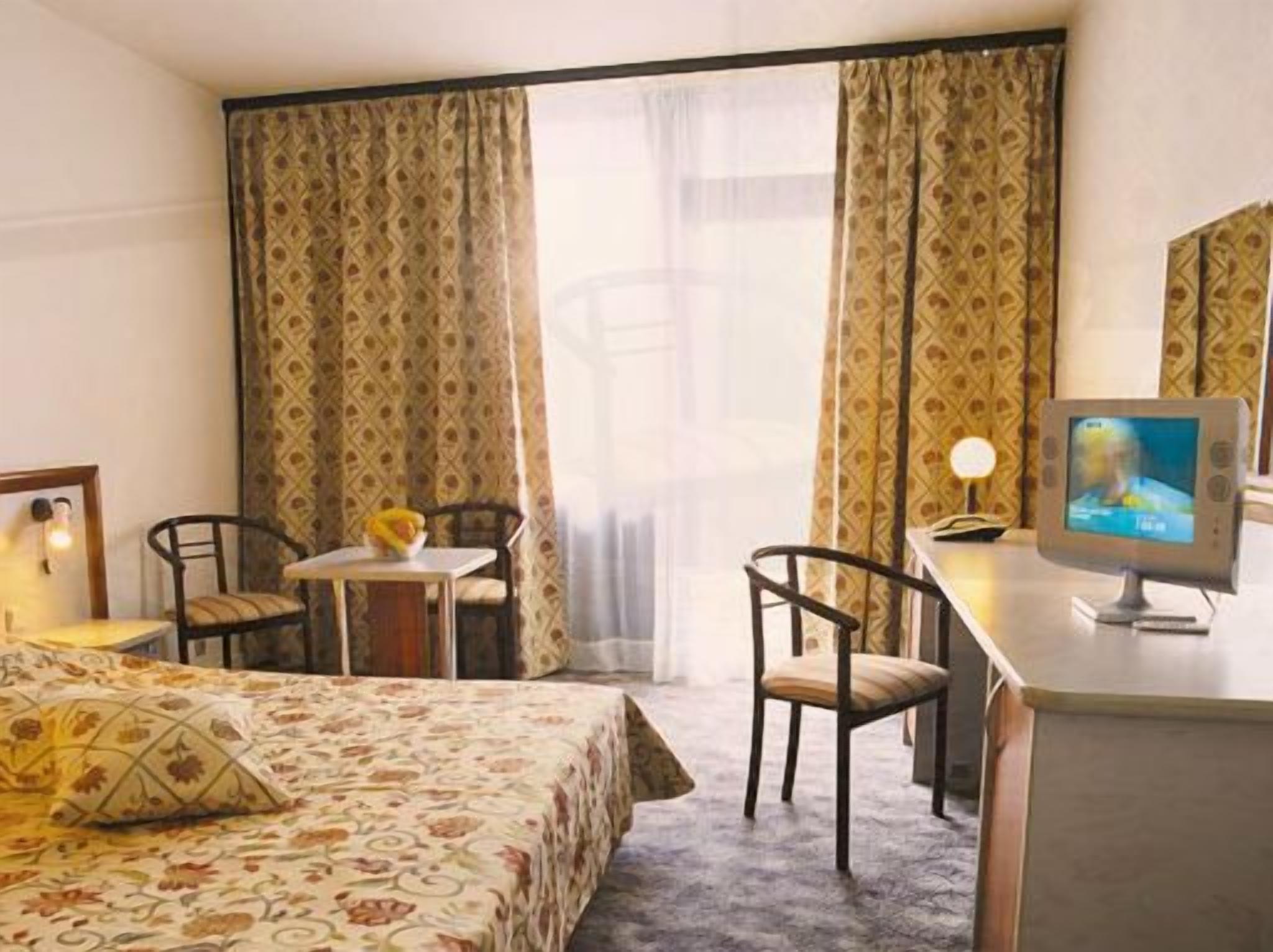 A hotel room with a bed, two chairs, a small table with a vase, and a television on a desk.