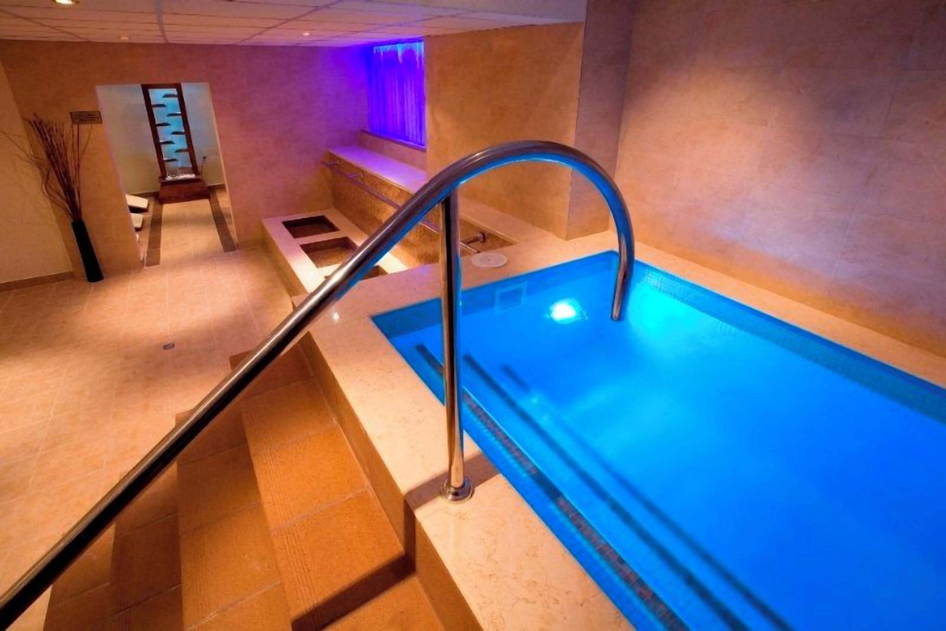 Una piscina con barandilla metálica, un banco y una pared iluminada de color morado.