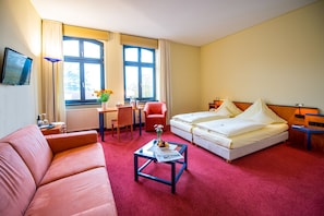 A hotel room with a red carpet, a sofa, a bed, a desk with a chair, and a TV mounted on the wall.