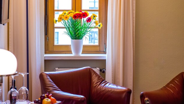 A room with a leather armchair, a table with a bottle of champagne, a vase of flowers, and a window with curtains. - Hotel Zum Ritter (Fulda)